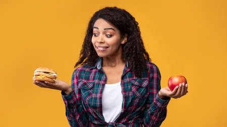 Junk Food. Hungry African American Lady Holding Burger And Apple Fruit. Funny Woman Looking At...