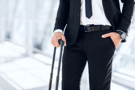 Wealth and success concept. Closeup of businessman standing by window in airport, holding luggage, copy space. Unrecognizable entrepreneur in black suit waiting for flight in modern airportの写真素材