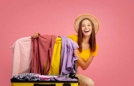 Smiling young woman tourist packing her luggage before vacation, showing her clothes to camera, pink...