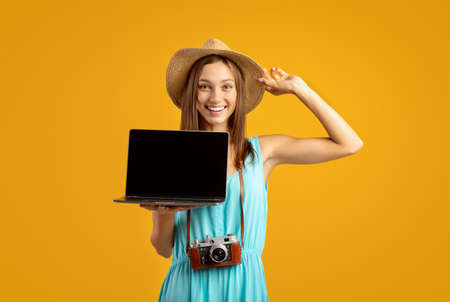 Cheerful young woman traveller holding laptop with empty screen over yellow studio background,...