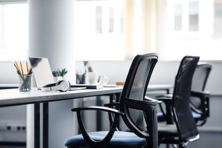 Modern gadgets in interior of coworking office during COVID-19 epidemic. Chairs and desk for support workers, laptops and headphones, cups and stationery in daylight on window background, free spaceの写真素材