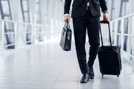 Unrecognizable entrepreneur in black suit walking by airport terminal, carrying luggage and bag, copy space. Rich businessman with case and suitcase going to business trip, croppedの写真素材