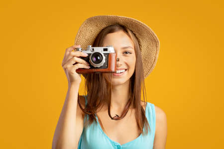 Portrait of cheerful woman photographer taking photos over yellow studio background. Young lady traveller wearing summer hat, shooting famous places, using professional camera, studio shotの写真素材