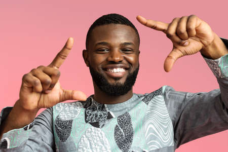 Playful black young man in colorful african costume showing photo frame with fingers on pink studio background, closeup. Happy african american guy demonstrating traditional blue and grey clothesの写真素材