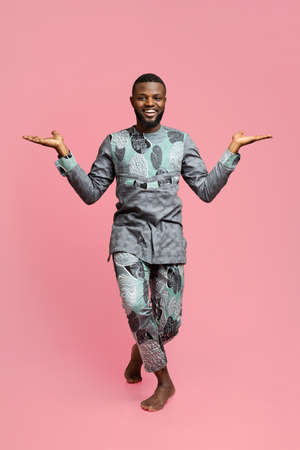 Cheerful african american man in traditional colorful costume dancing over pink studio background,...