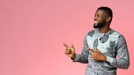 Excited black guy in african costume pointing at free space for advertisement, pink background,...