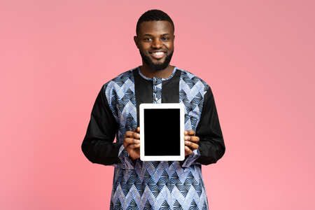 Cheerful black guy holding digital tablet with blank screen, wearing traditional african costume....