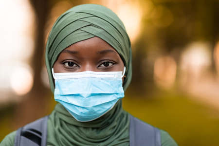 Outdoor portrait of african muslim woman in headscarf wearing protective medical mask on face for coronavirus protection, standing on street and looking at camera, closeup shot with selective focusの写真素材
