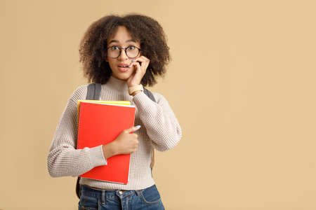 Excited human emotions. Amazed african american lady with glasses, open mouth, presses her hand to face, holds notebooks and back to school after coronavirus quarantine, isolated on yellow backgroundの写真素材