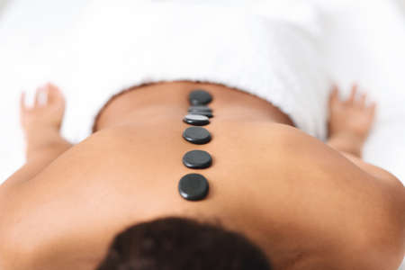 Black stones on naked unrecognizable black lady back, spa interior, closeup. Cropped of african american good looking woman attending spa salon, getting healing hot stone massageの写真素材
