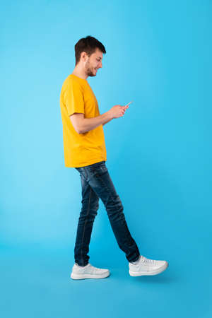 Communication, Technology And Lifestyle Concept. Full length portrait of handsome smiling young man using mobile phone, walking isolated over blue studio background, vertical shotの写真素材