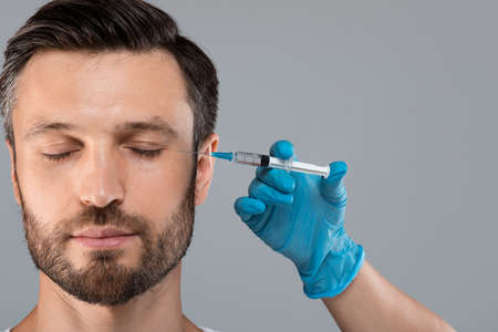 Middle-aged bearded man getting anti-wrinkle injection in eye zone over grey studio background. Cosmetician injecting filler for handsome man in beauty salon, copy space. Male cosmetology conceptの写真素材
