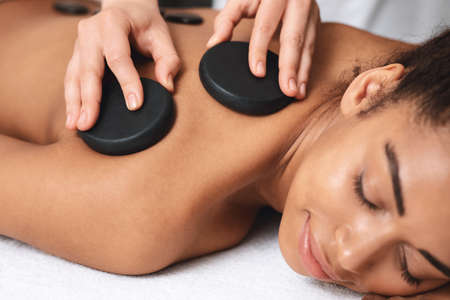 Day at spa. Closeup of young black lady with closed eyes having healing hot stone massage at beauty...