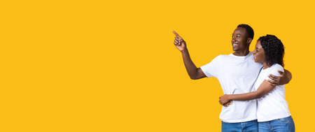 Cheerful black guy showing his girlfriend copy space on yellow studio background. African american lovers hugging and looking at empty space for your advertisement or text, panoramaの写真素材