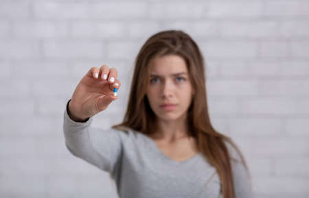Depressed young lady showing antidepressant or sedative tablet against white brick wall, selective...