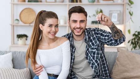 Buying or renting real estate. Happy millennial husband and wife sitting on sofa and looks at camera, man showing keys to new apartment in interior of modern living room, free spaceの写真素材