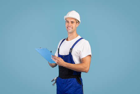 Repair And Renovation. Smiling handyman in hard hat helmet posing over blue background in studio, holding clipboard, taking notes, positive maintenance employee ready to work, looking at cameraの写真素材