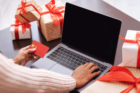 Online Shopping Concept. Over the shoulder view of black female hands holding red credit card and using laptop with empty blank screen with mock up, ordering presents and making paymentの写真素材