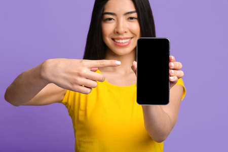 Positive Asian Lady Pointing On Blank Black Smartphone Screen, Showing Free Space For Advertisement, Posing Over Purple Background In Studio, Mockup Image, Creative Template For App Or Website Designの写真素材