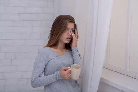 Unhappy young lady standing near window with cup of coffee and crying, indoors. Depressed millennial woman feeling upset, stressed or lonely, suffering from mood swings or PTSDの写真素材