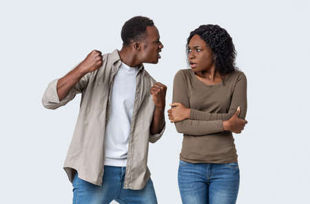Domestic violence and abuse concept. Young african american mad man threatening his woman with his fist, gray studio background. Furious black guy showing fists his scared girlfriend or wifeの写真素材