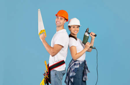 Repair Team. Happy Couple In Hard Hats Posing With Hand Saw And Drill Over Blue Background In...