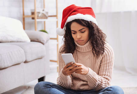 Frustrated african american woman using cellphone sitting on the floor in living room. Confused...