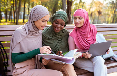 Three Muslim Students Women Learning Together Reading Books And Using Computer Sitting On Bench In University Campus Park Outdoors. Education For Modern Arabic Ladies, Online Study Conceptの写真素材