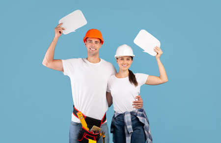 Happy Young Couple In Hard Hats Holding Empty Speech Bubbles In Hands Posing Over Blue Background In Studio, Advertising Repair Services, Having Home Renovation, Free Copy Space For Your Textの写真素材
