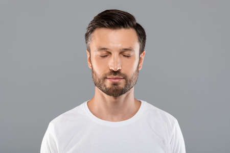 Middle-aged bearded man with closed eyes posing over grey studio background, free space. Handsome man demonstrating his healthy good looking skin, advertisement for male cosmetologyの写真素材