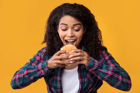 Junk Food. Closeup Portrait Of Funny Hungry African American Woman Eating Burger. Black Lady With Curly Hair Holding And Taking Bite Of Tasty Sandwich, Isolated Over Orange Studio Background, Bannerの写真素材