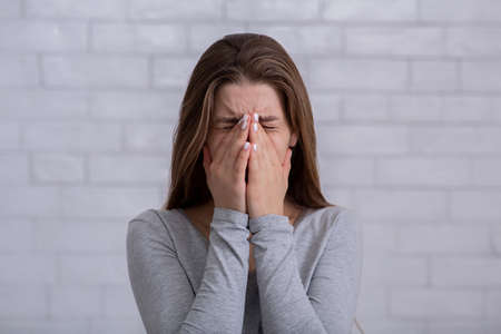 Depressed millennial woman covering her face and crying against white brick wall. Sad young lady...