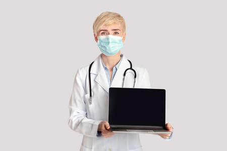 Online application for visit to therapist. Serious adult woman in white coat with stethoscope and protective mask shows laptop with blank screen, isolated on gray background, studio shot, cut outの写真素材