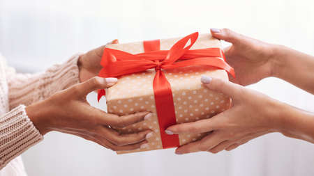 Holiday Celebration Concept. Closeup on female hands holding gift with red bow and giving present box to african american woman, greeting with Birthday, Christmas and New year at homeの写真素材