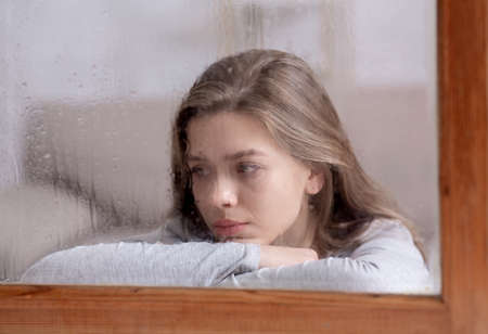 Unhappy young lady looking out window with drops of rain, feeling sad or melancholic. Emotional...