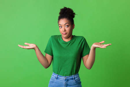 Who Knows. Portrait of Funny Young Black Woman Shrugging Shoulders Looking At Camera Posing Over Green Background. Studio Shot. Puzzled lady is clueless. Im sorry, exuse me, cant decideの写真素材