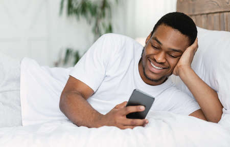 Smiling African Guy Using Cellphone Browsing Internet And Texting On Phone Lying In Bed At Home....