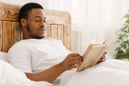 Black Man Enjoying Reading Book In The Morning Lying In Comfortable Bed At Home. Bookworm Guy Reads Longread Novel Relaxing On Weekend In Bedroom. Read More Concept. Selective Focusの写真素材