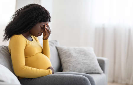 Young pregnant black woman suffering from headache or migraine, feeling sick, sitting on sofa at home, free space. Sad african american expecting lady touching her forehead and big tummy, home aloneの写真素材
