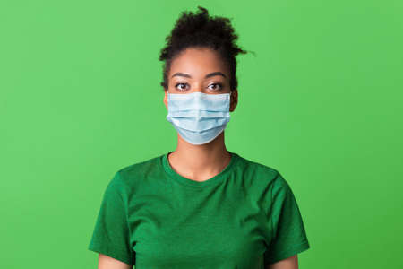 Health Care And Flu Concept. Closeup front portrait of african american young woman wearing surgical mask isolated on green studio wall background. Female patient looking at camera, bannerの写真素材