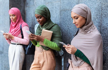 Modern Arab Ladies Using Smartphones Mobile Apps Texting And Networking In Social Media Standing Over Gray Wall Outside. Female Muslim Students With Cellphones Using Applicationsの写真素材
