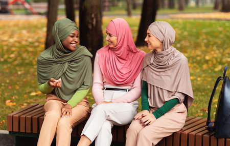 Three Muslim Ladies In Hijab Headscarf Talking Sitting On Bench In Park Outdoors. Female Friendship,...