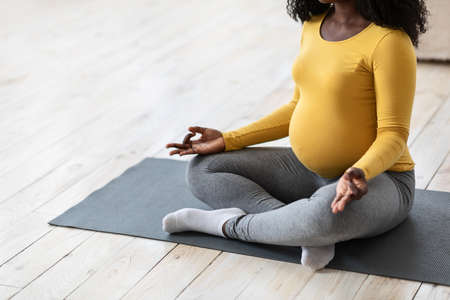 Unrecognizable pregnant black lady meditating at home or yoga studio, copy space. Cropped of african expecting woman practicing yoga, sitting on floor. Sport and healthy lifestyle during pregnancyの写真素材