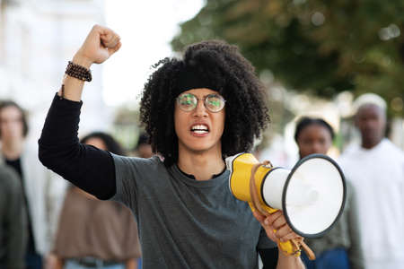 Mixed race guy with megaphone leading international group of protestors on the street, raising fist up, chanting slogans. Leader of strikers with loudspeaker yelling demands to governmentの写真素材