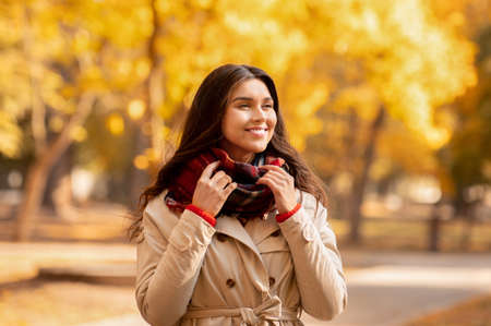 Beautiful millennial lady in coat and warm scarf enjoying colorful change of seasons in autumn park....