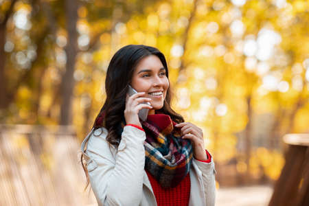 Pretty millennial woman having friendly conversation on cellphone at autumn park. Portrait of smiling young lady with mobile device on walk outside on sunny fall dayの写真素材