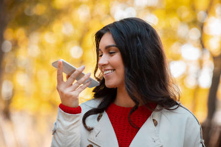 Beautiful young woman speaking on smartphone at park on warm autumn day. Pretty millennial lady...
