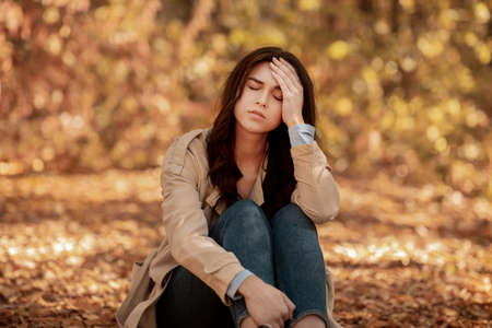 Tired or depressed young woman touching her head at autumn park. Millennial lady suffering from terrible headache, feeling lonely, stressed or unhappy, having problemの写真素材