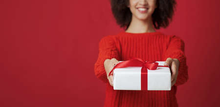 portrait of smiling african american female in sweater gives box with gift, isolated on red background, empty space, close up, panoramaの写真素材