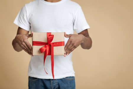 Surprise for Christmas, New Year and family party. African american man in white t-shirt holding box with gift in package with ribbon isolated on light background, cropped, studio shot, free spaceの写真素材